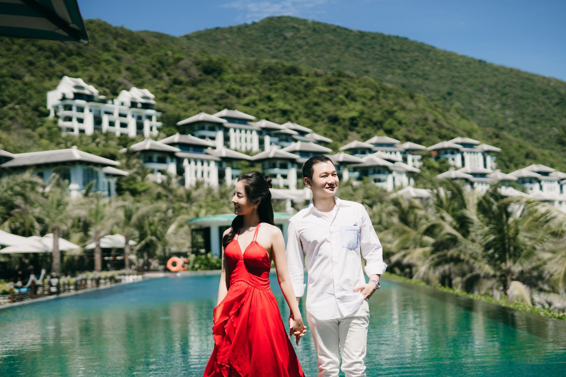 Couple walking by a turquoise resort pool with hillside villas in the background
