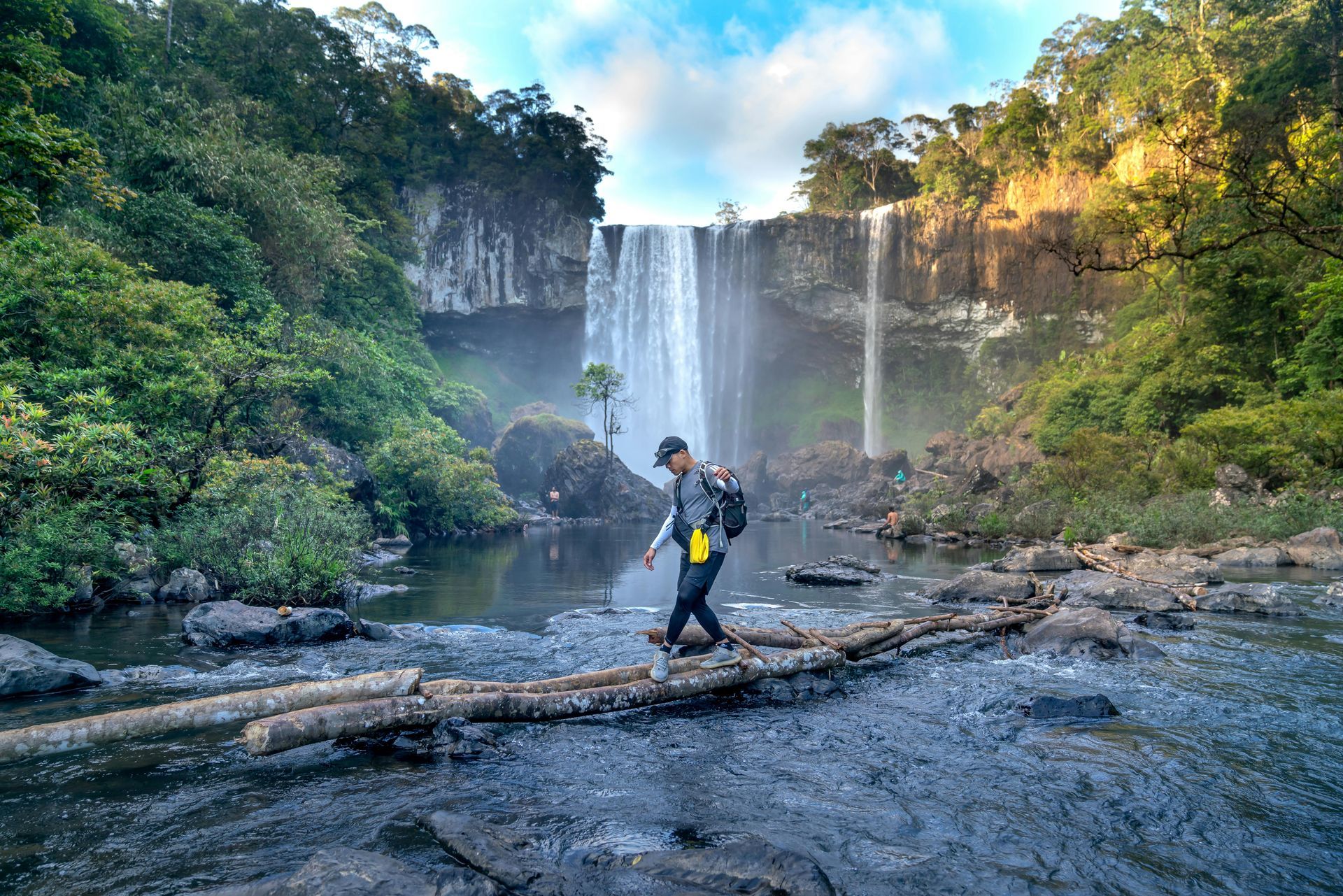 Person in a helmet crossing a log in front of a tall waterfall in a lush forest