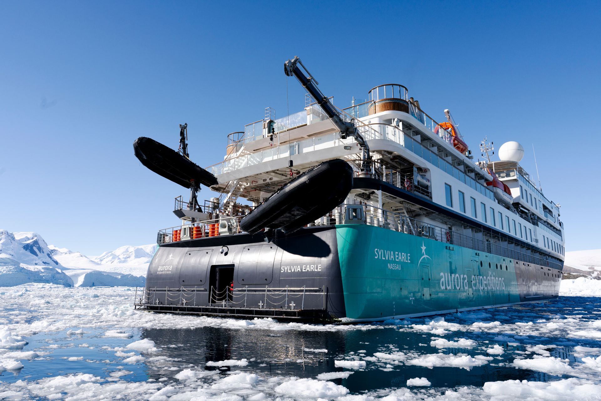Large cruise ship navigating icy polar waters near snow-covered mountains under a clear blue sky