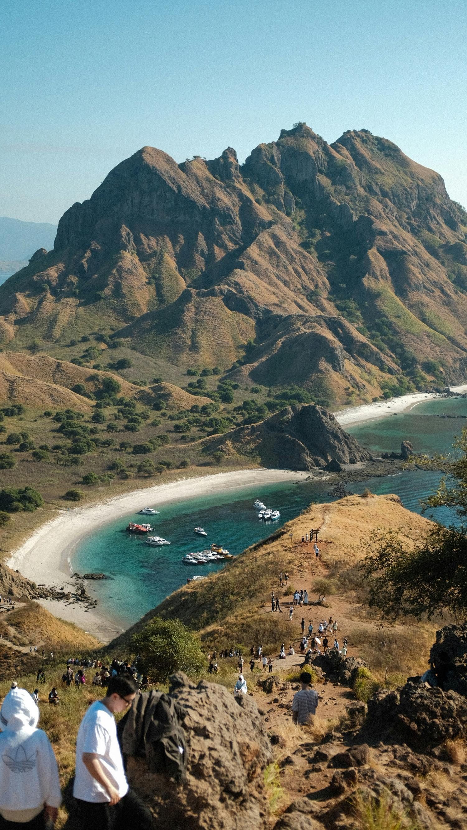 Coastal bay with turquoise water, small boats, rocky mountain, and hikers on a sunlit trail