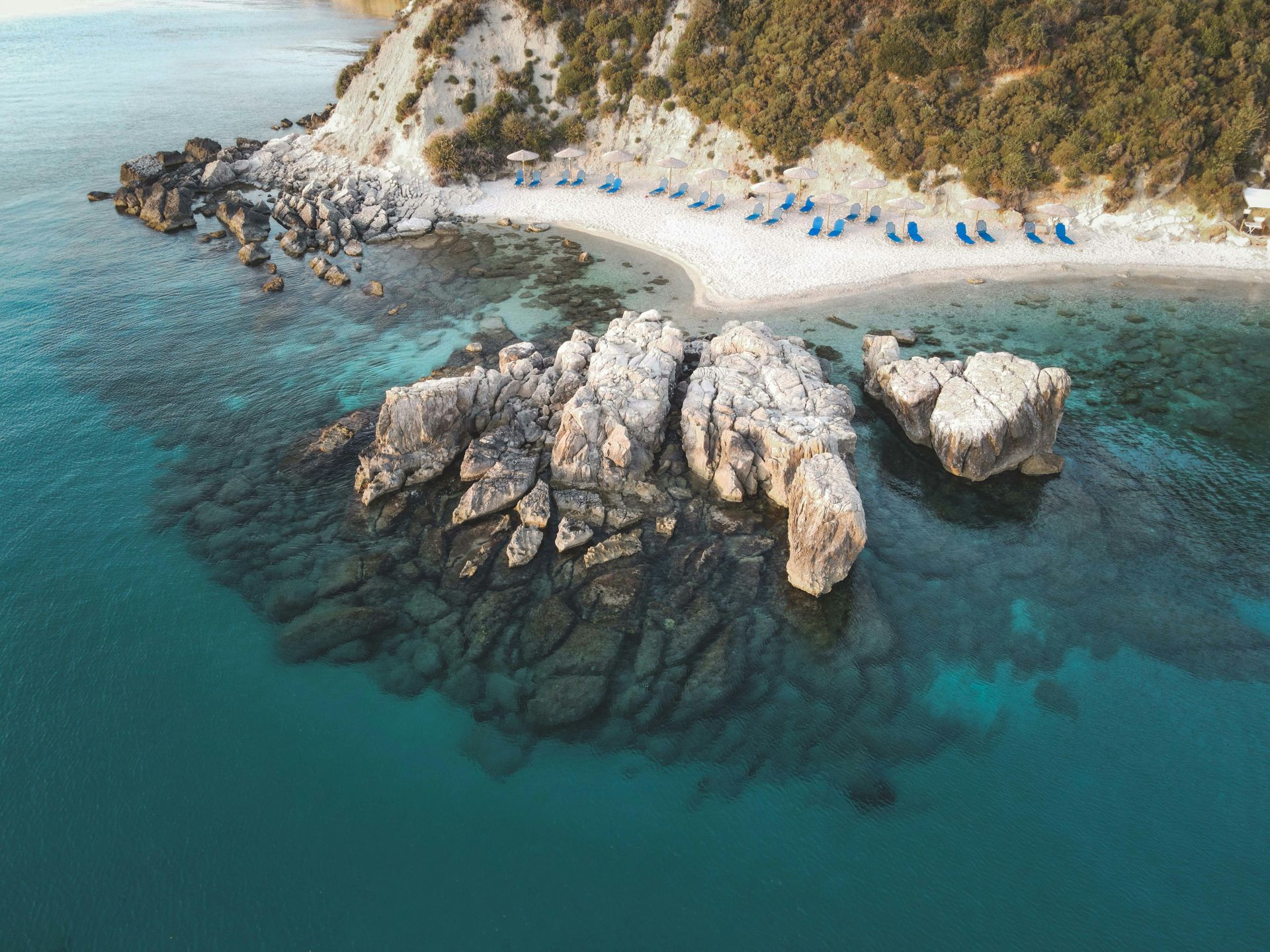 Aerial view of a rocky turquoise cove with a small sandy beach and blue umbrellas along the shore