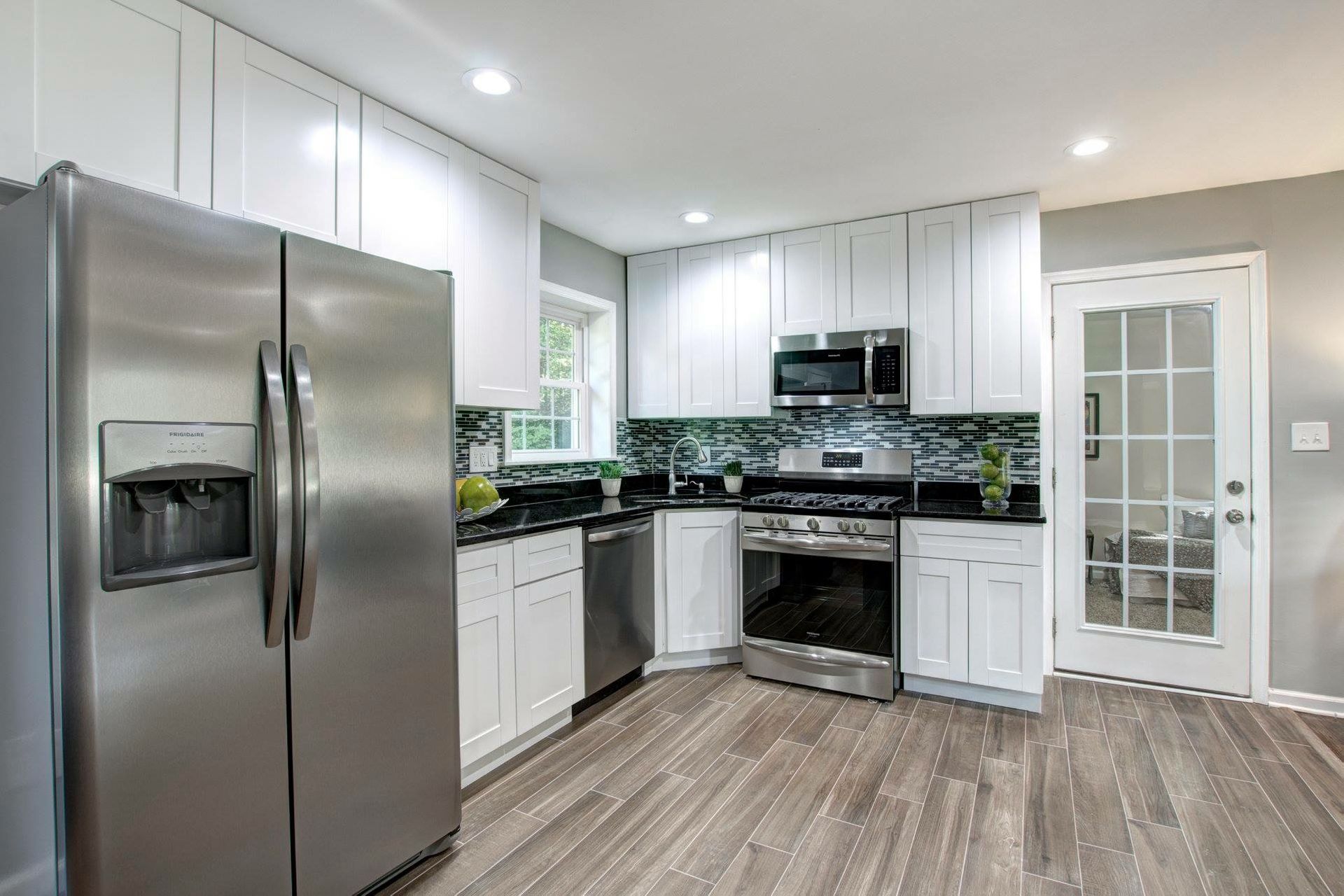 Bright modern kitchen with stainless steel fridge, black countertops, white cabinets, and a glass door.