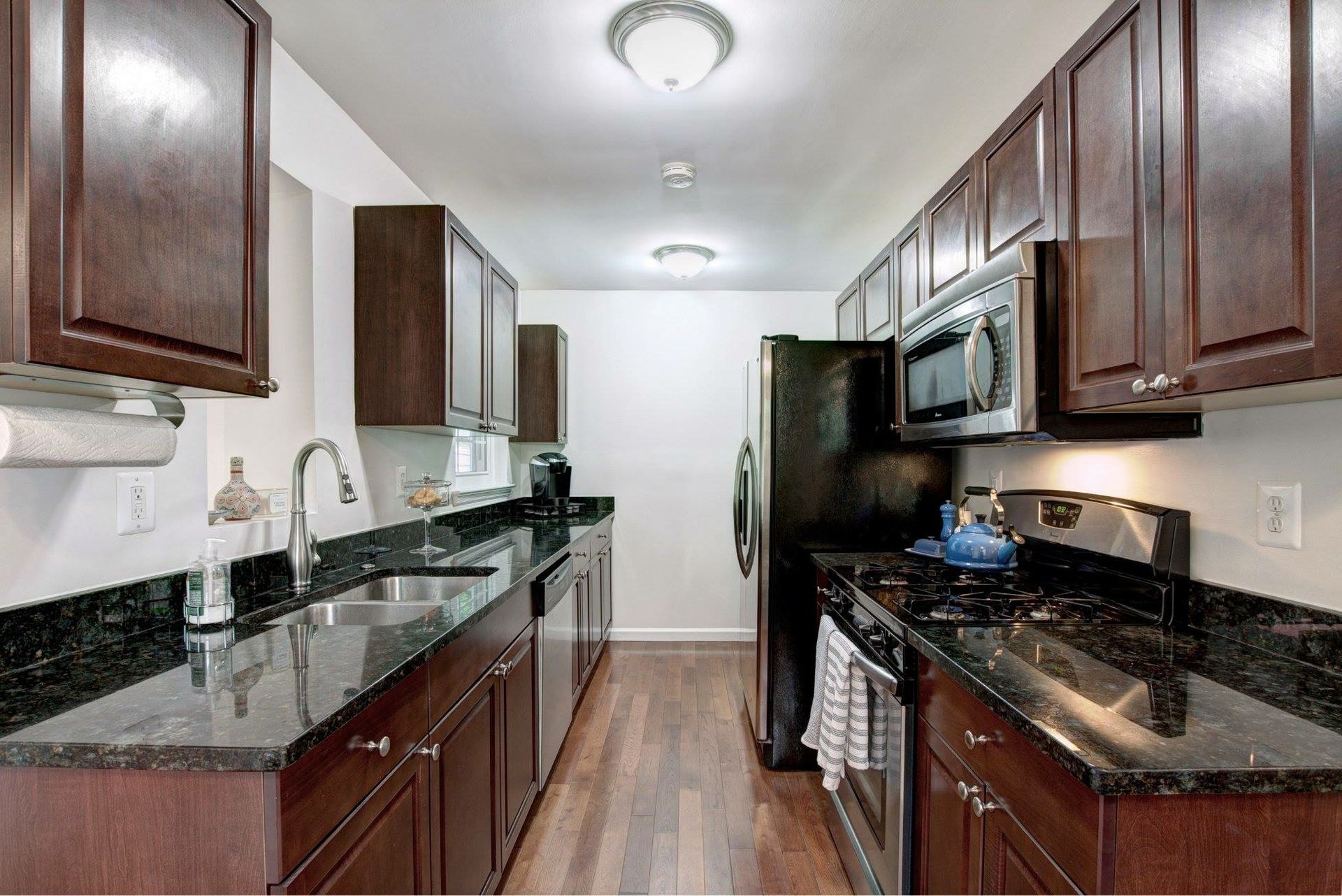 Narrow modern kitchen with dark wood cabinets, black granite counters, and stainless steel appliances.