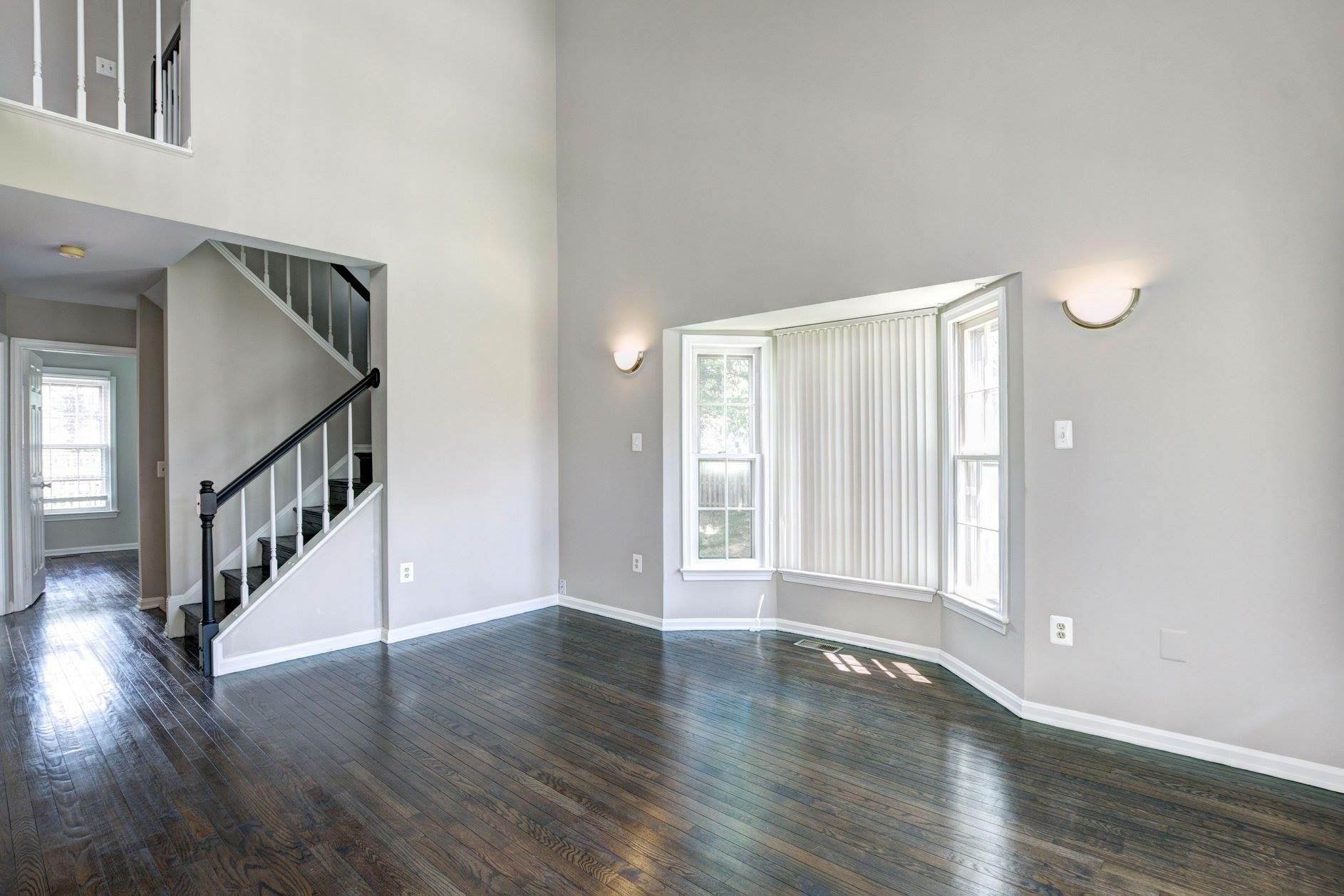 Empty living room with dark hardwood floors, gray walls, bay window, and staircase