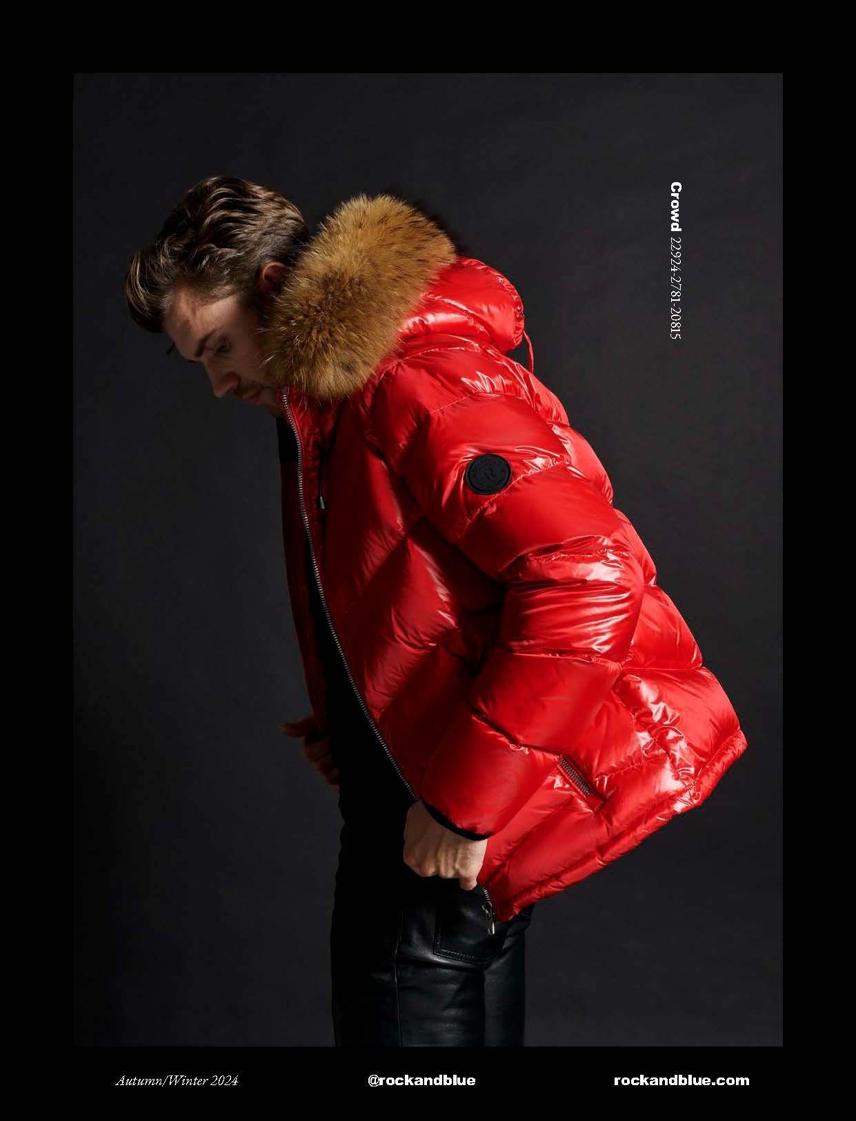Mann in einer glänzenden roten Daunenjacke mit pelzgefütterter Kapuze, Hände in den Taschen, vor dunklem Hintergrund.