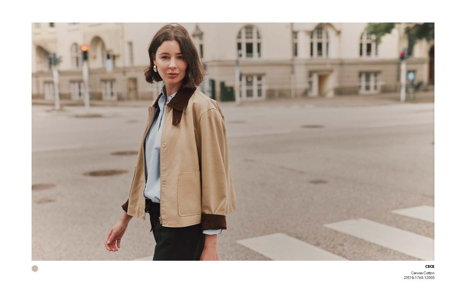 Eine Frau überquert eine Straße. Sie trägt eine beige Jacke mit braunem Kragen. Im Hintergrund sind Gebäude zu sehen.