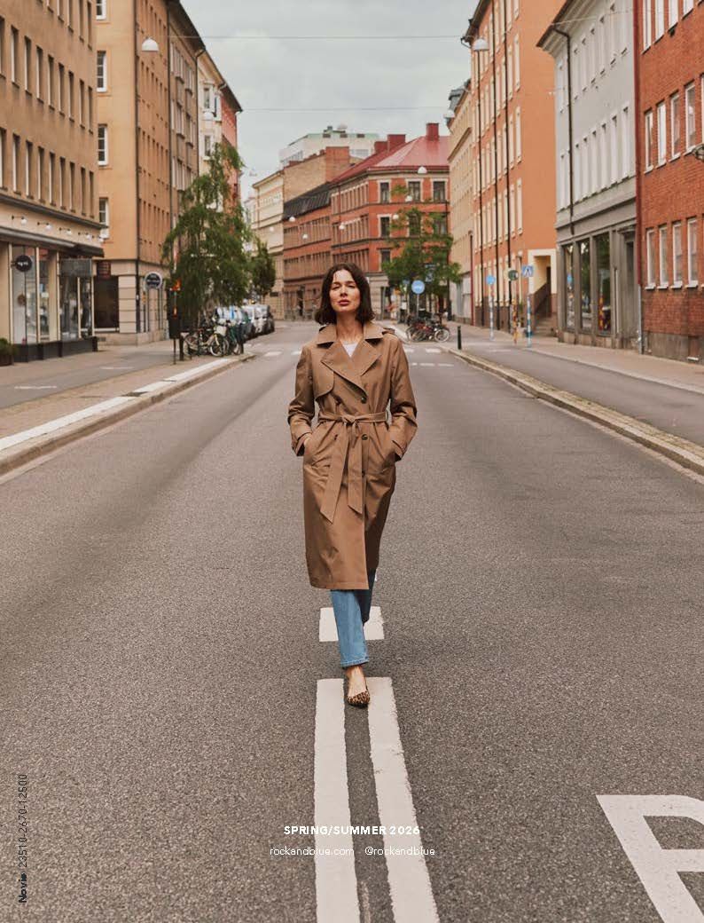 Eine Frau in einem beigen Trenchcoat geht eine Straße in der Stadt entlang. Gebäude säumen die Straßenränder.