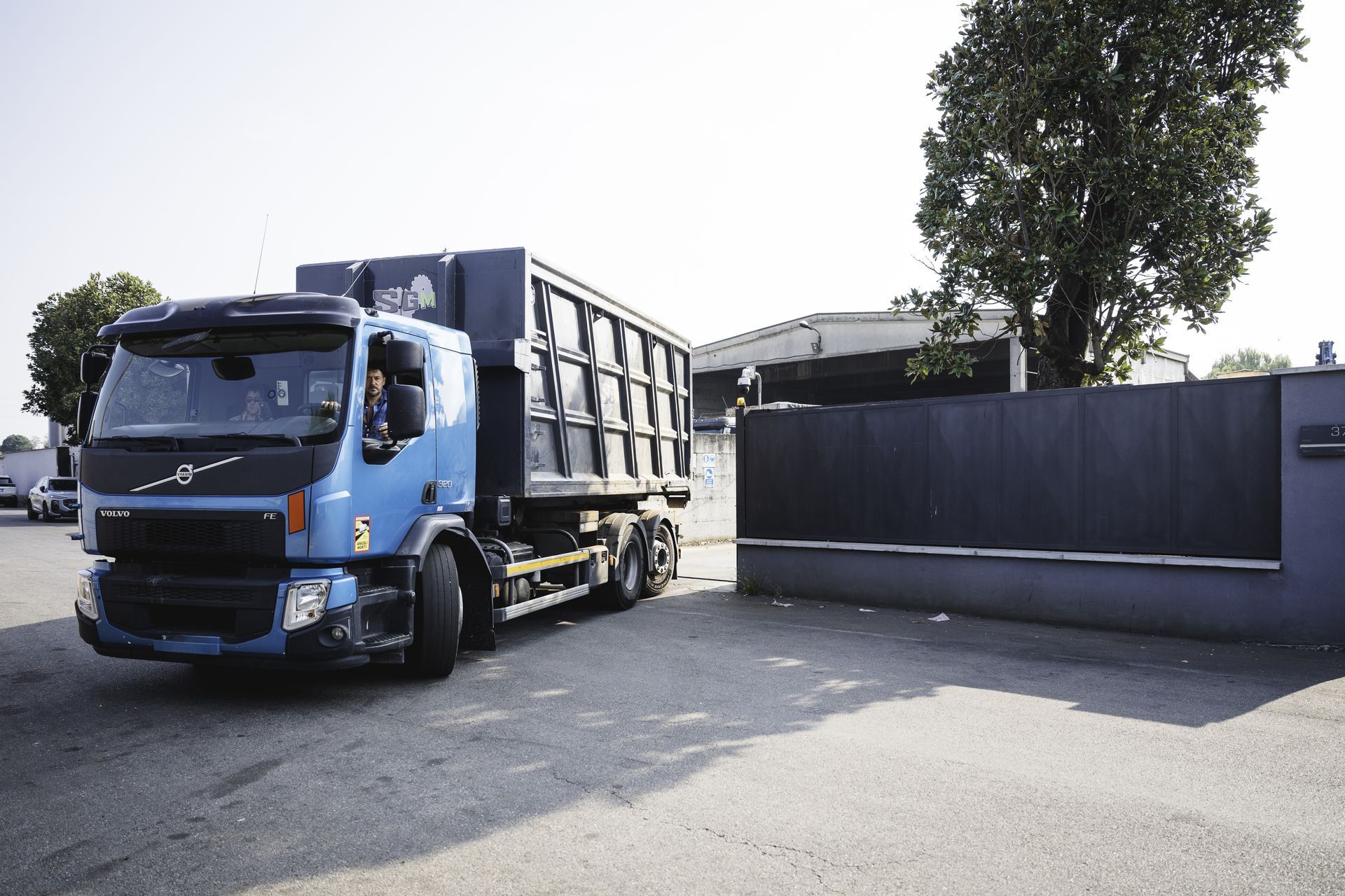 Un camion ribaltabile arancione parcheggiato accanto all'area di carico di un magazzino, con un grande serbatoio bianco davanti.