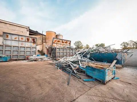 Area industriale danneggiata con una struttura metallica blu crollata e detriti davanti a un magazzino.