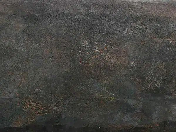Superficie scura, screziata di grigio-marrone, dalla consistenza ruvida che ricorda la pietra o il cemento erosi dagli agenti atmosferici.