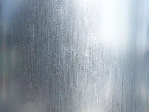 Primo piano di una superficie metallica argentata spazzolata con delicate striature verticali e riflessi luminosi.