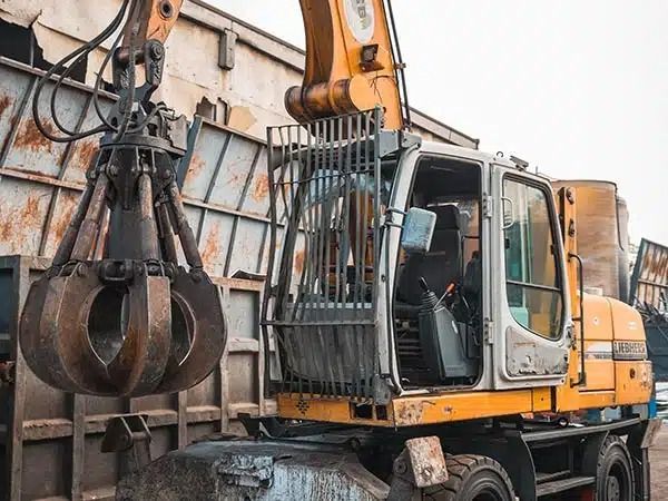 Escavatore arancione con pinza, parcheggiato accanto a un muro di rottami metallici.