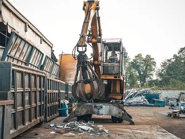 Escavatore con pinza che solleva rottami metallici in un'area industriale ingombro di oggetti, accanto a un magazzino.