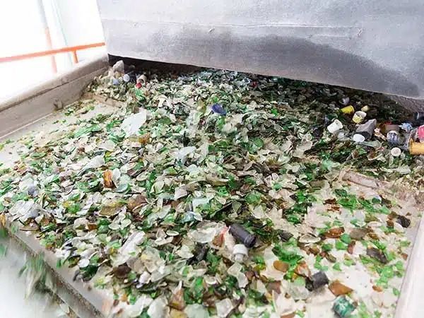 Cumulo di bottiglie e lattine miste riciclabili sul pavimento vicino a una superficie metallica e a una ringhiera.