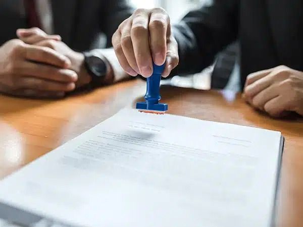 Mani che timbrano un documento su un tavolo di legno con un timbro blu e sullo sfondo fogli di carta sfocati.
