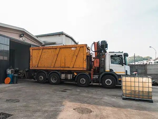 Un camion ribaltabile arancione parcheggiato accanto all'area di carico di un magazzino, con un grande serbatoio bianco davanti.