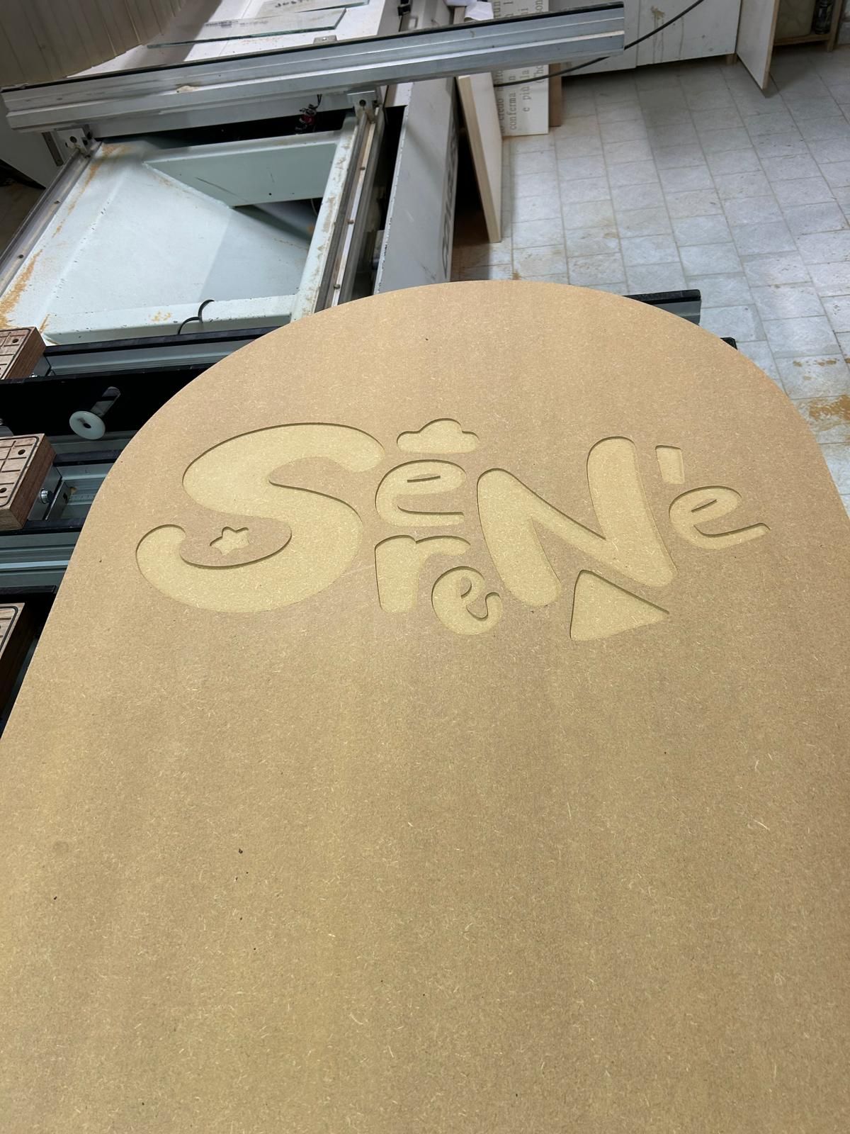 Scritta “SENE” tagliata al laser su un pannello di legno color cuoio in un'officina.