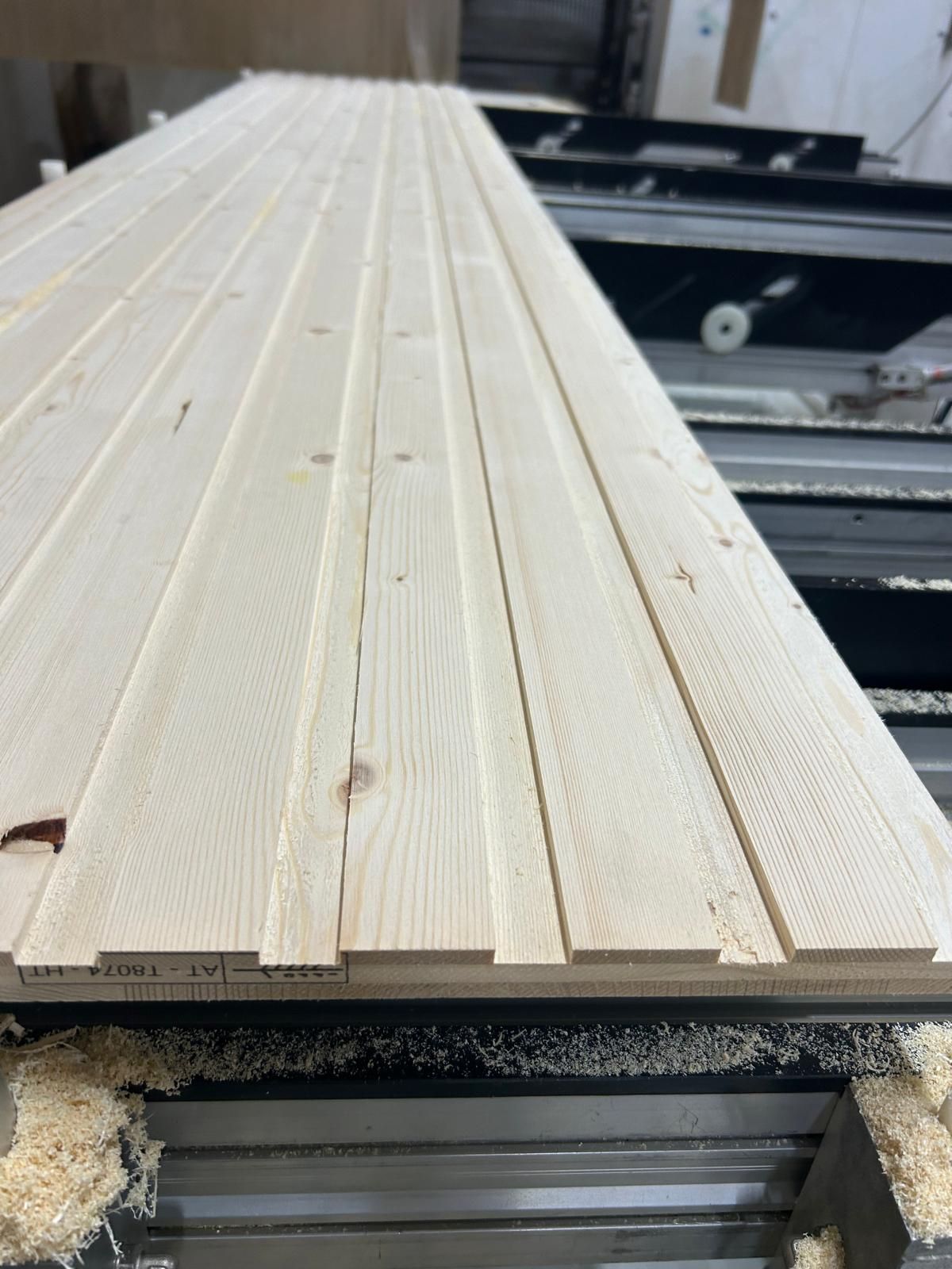 Pila di assi di pino chiaro sul nastro trasportatore di una macchina per la lavorazione del legno, con segatura visibile al di sotto.
