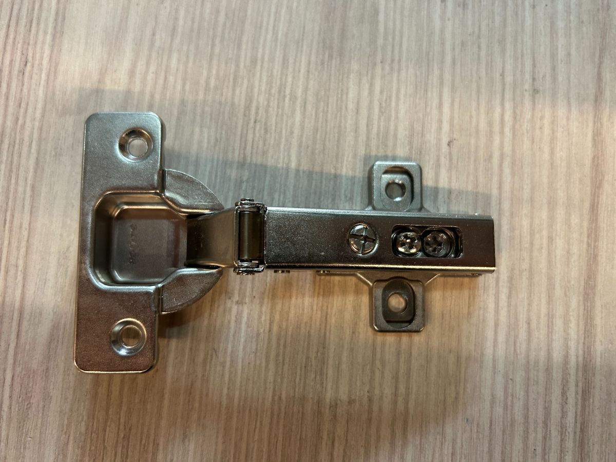 Cerniera per mobile in argento montata su una superficie in legno chiaro.