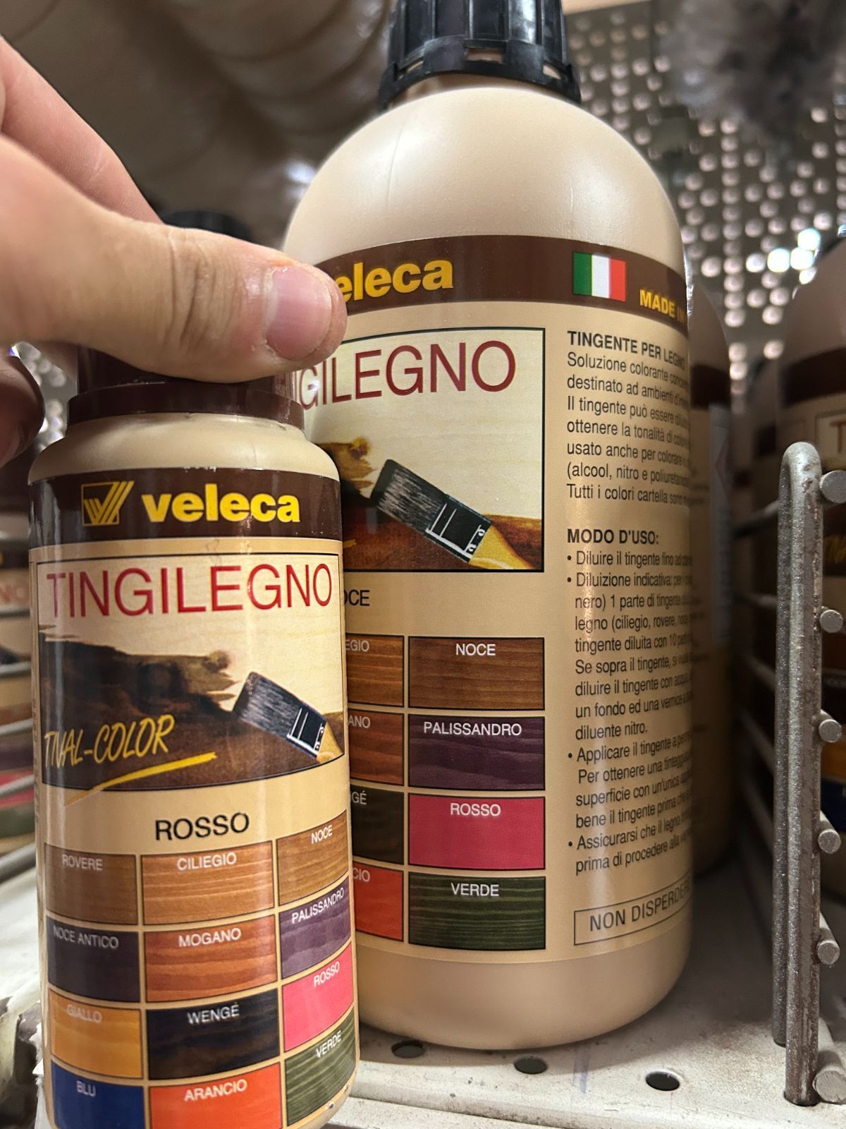 Una mano che tiene due barattoli beige di stucco per legno Veleca TingiLegno su uno scaffale di un negozio.