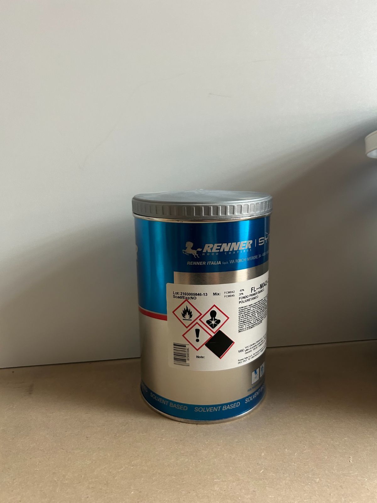 Contenitore blu per prodotti chimici con etichette di pericolo e coperchio argentato su un tavolo.