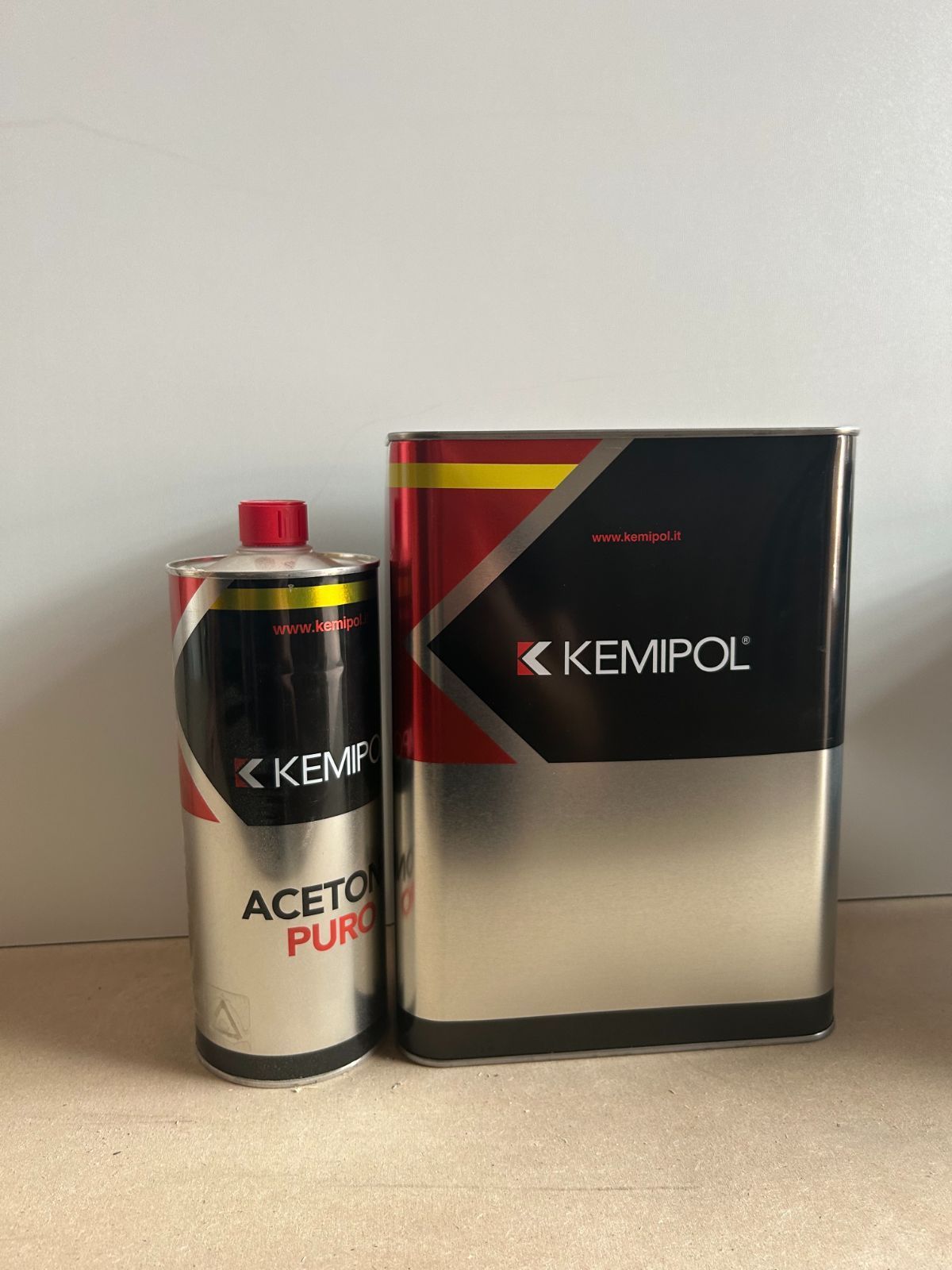 Una lattina di diluente acetone Kemipol accanto a una lattina di metallo nera Kemipol su una superficie piana.