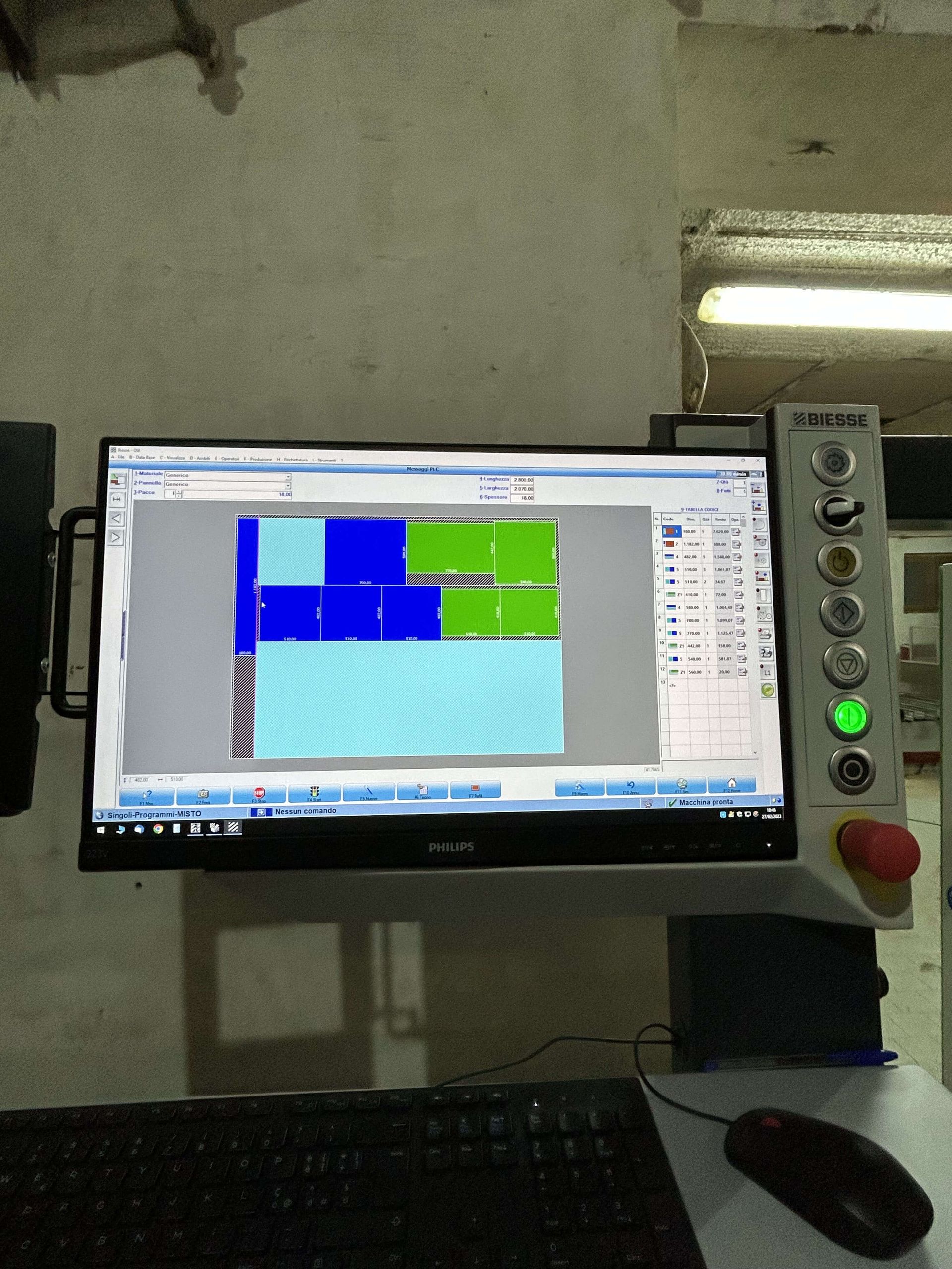 Monitor di controllo industriale con blocchi blu e verdi su una console di macchina, accanto a pulsanti illuminati e una tastiera.