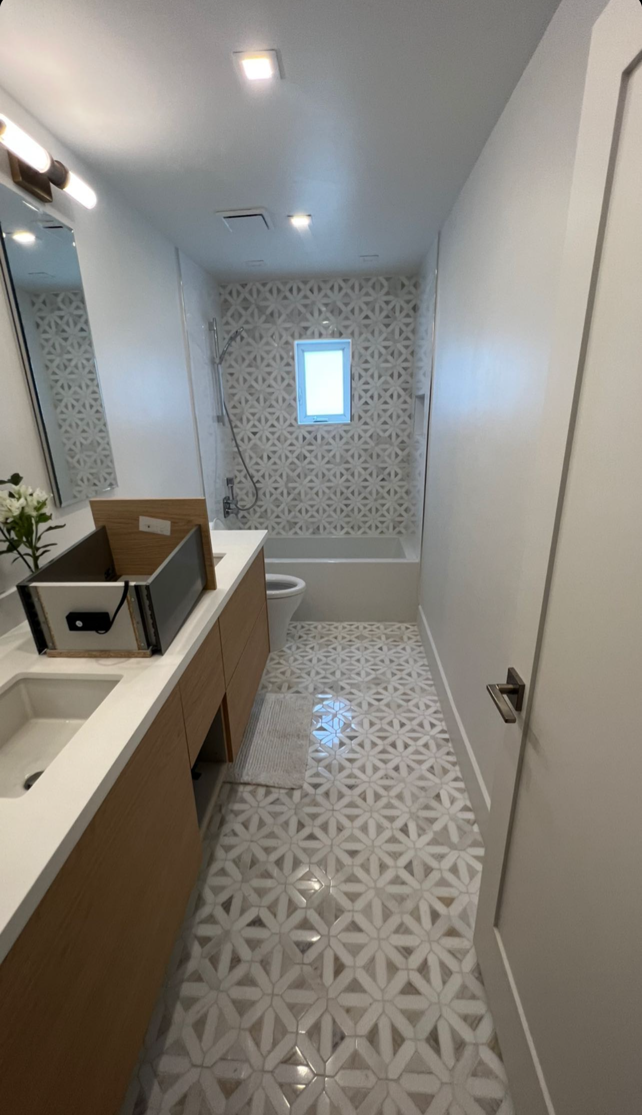Narrow modern bathroom with patterned floor tiles, white walls, vanity sink, mirror, and small window.