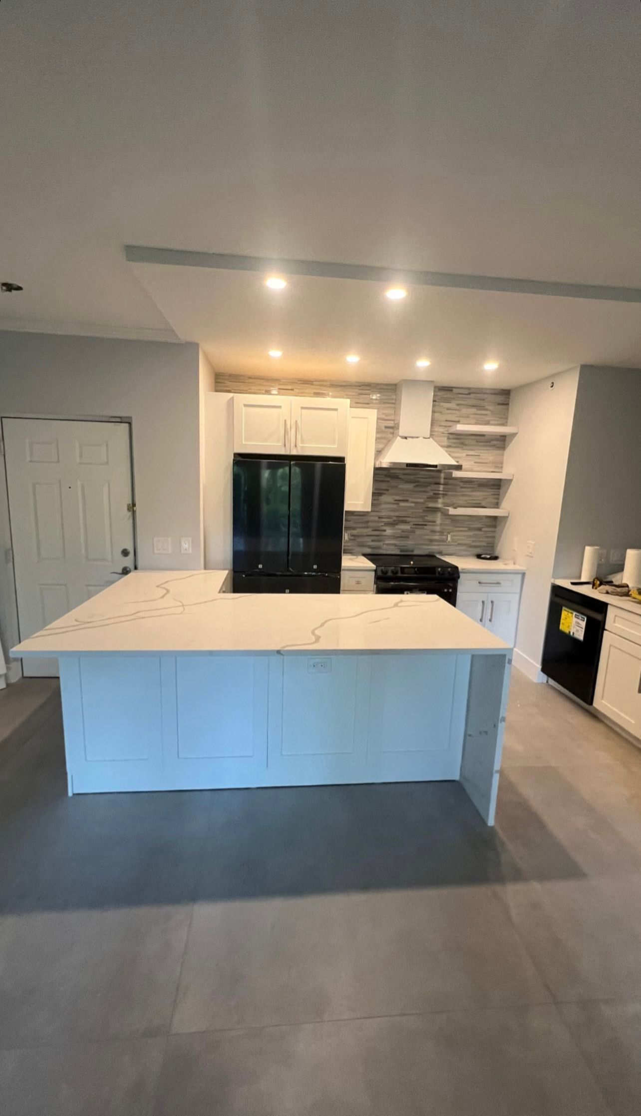 Modern kitchen with white island, dark fridge, open shelving, and grey floor tiles