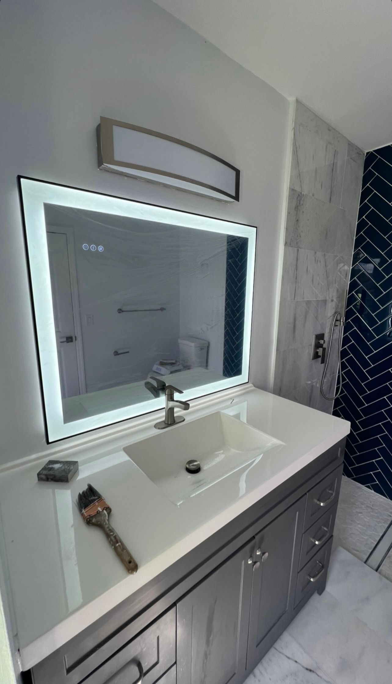 Bathroom vanity with illuminated mirror, white countertop, and gray cabinets beside a tiled shower.