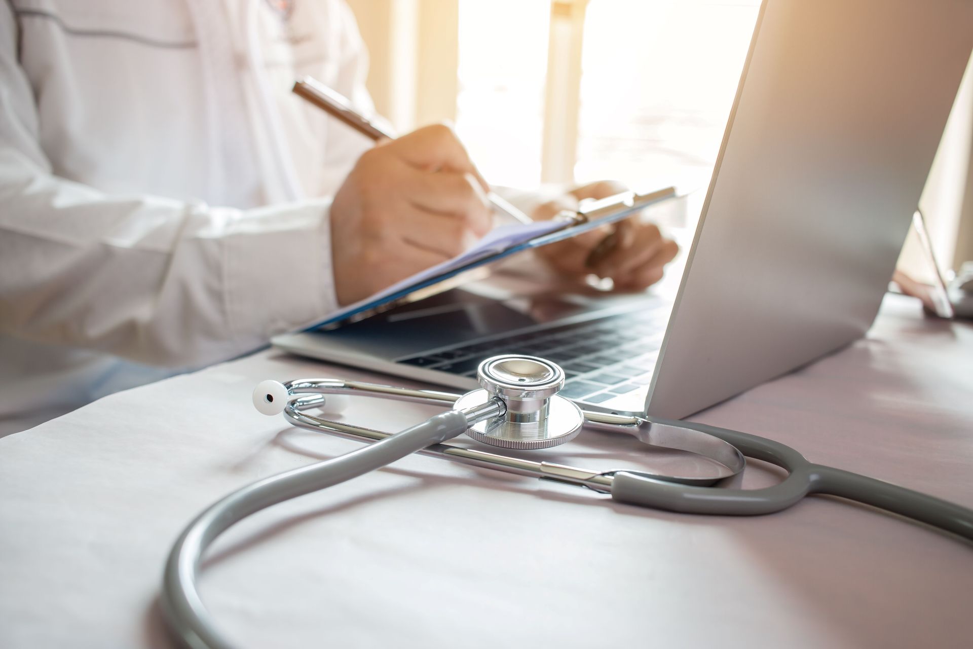 Doctor writing on clipboard beside a laptop with a stethoscope on a desk