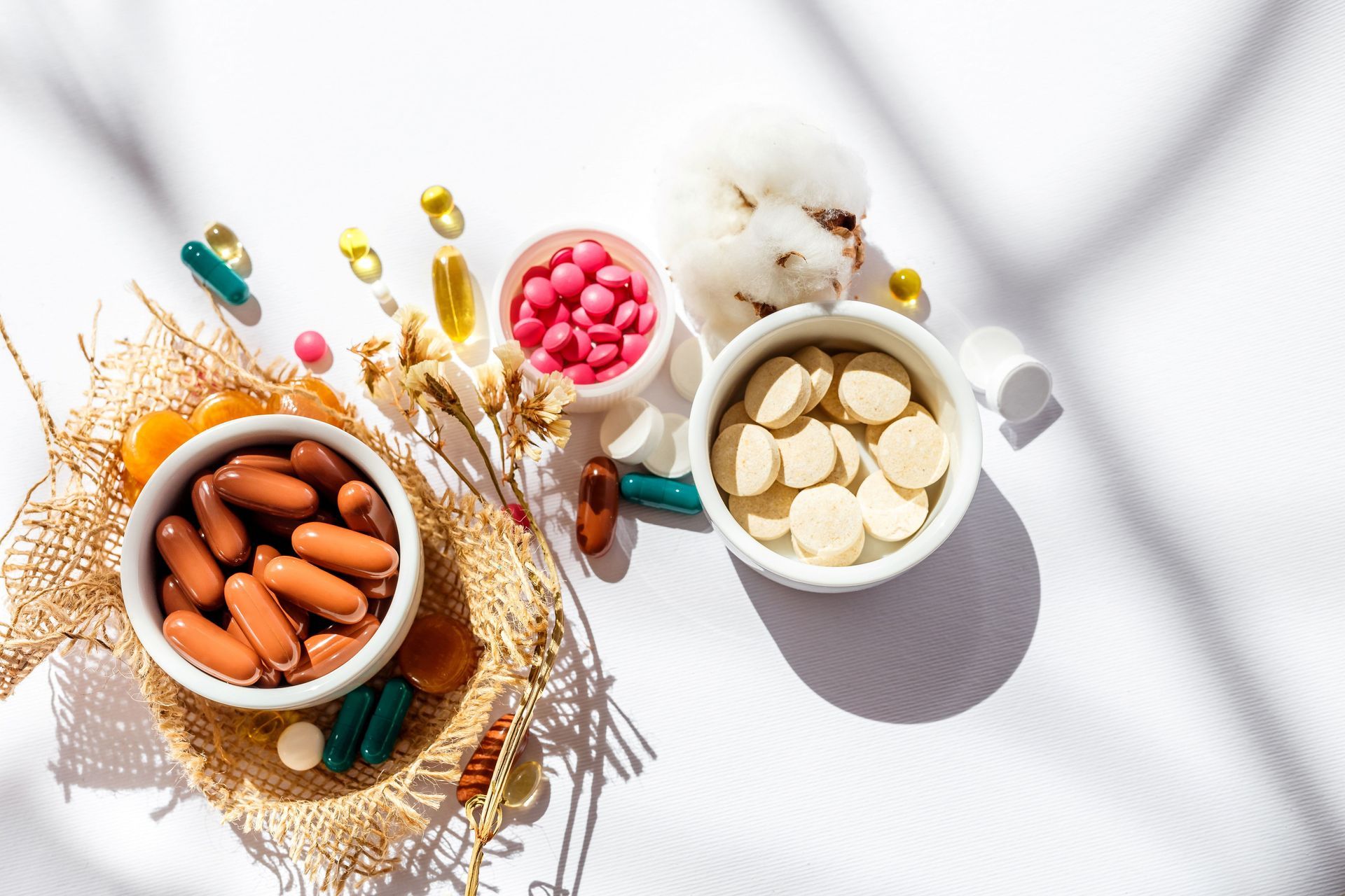Vitamin capsules and pills in bowls on a white surface with spilled supplements and cotton flowers