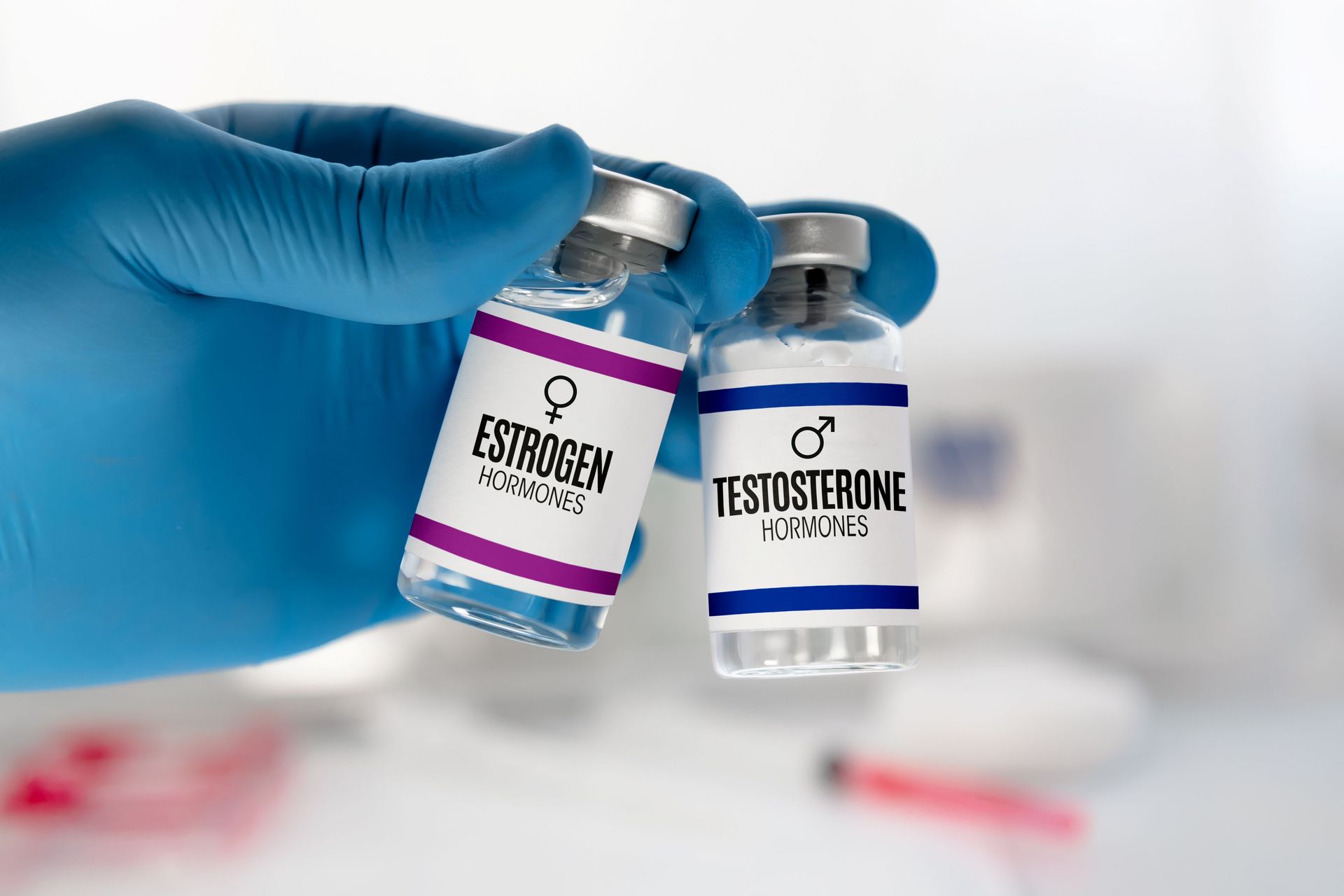 Gloved hand holding two small vaccine vials labeled estradiol and testosterone