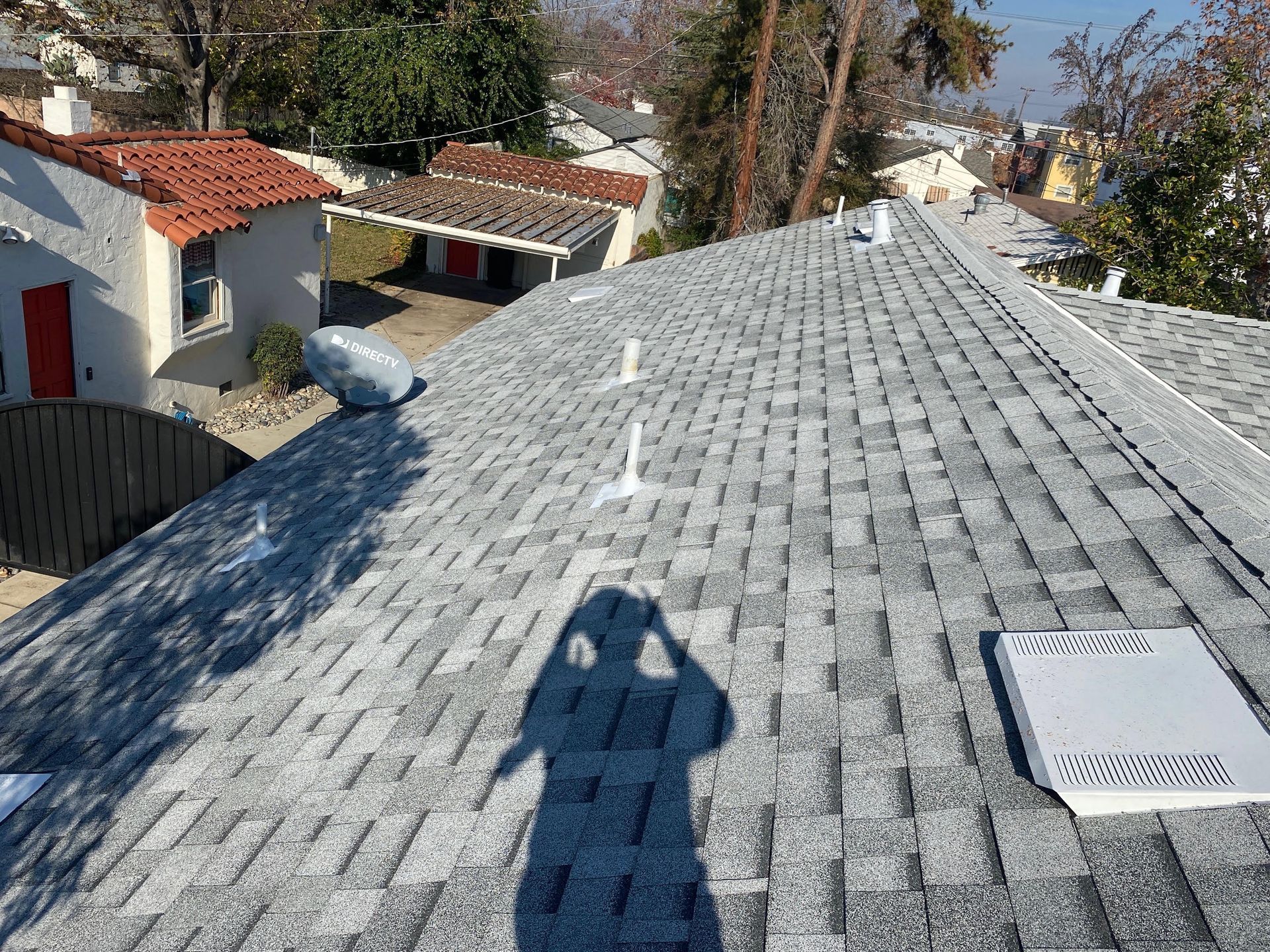 Roofer’s shadow on a gray shingle roof with a skylight, overlooking a residential neighborhood.