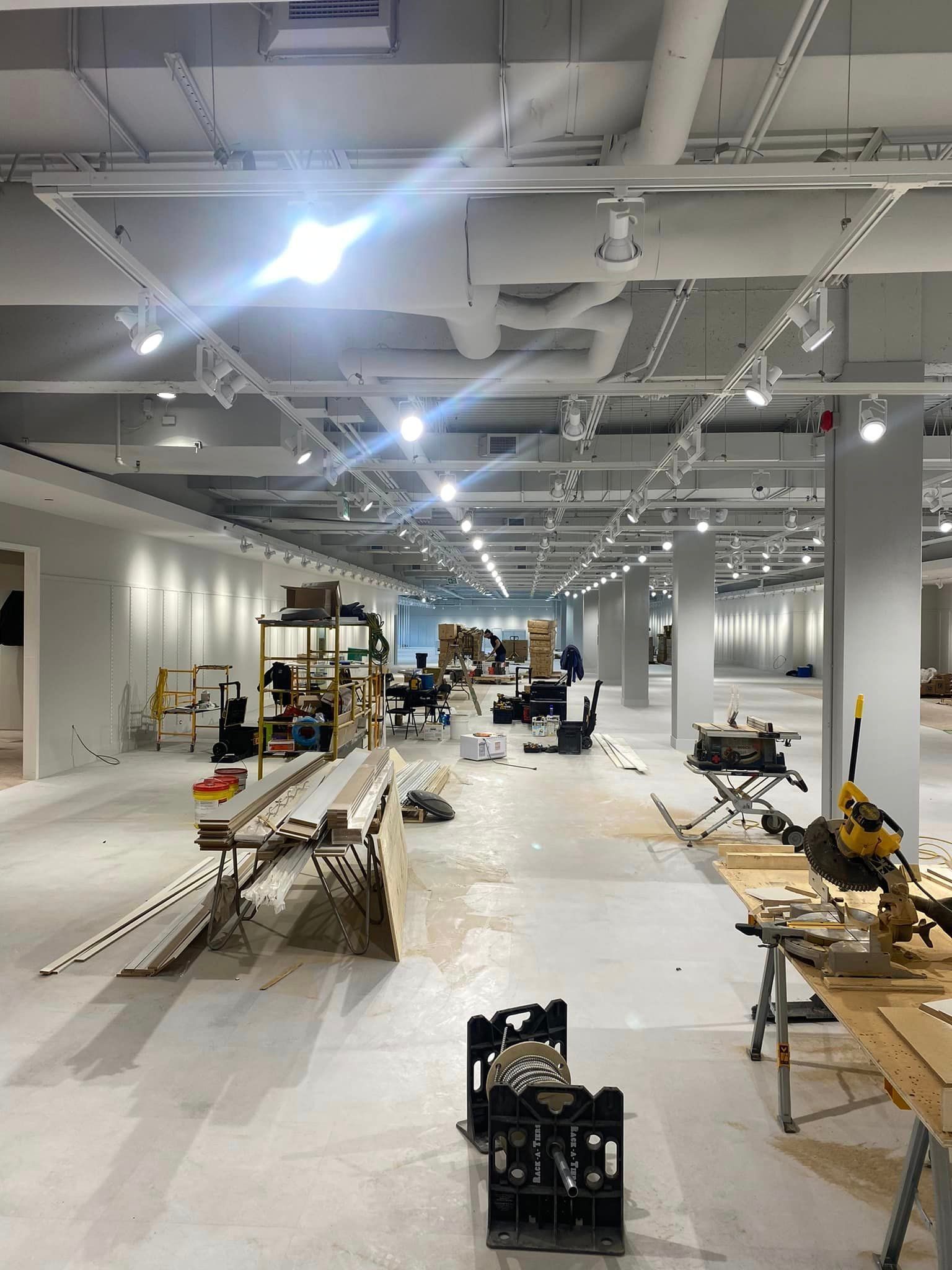 Grand espace de travail ouvert en construction avec des ouvriers, des outils, des tables, et des luminaires et conduits apparents au plafond.