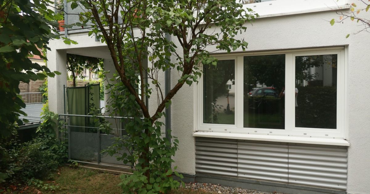 Modern home exterior with white facade, large windows, small tree in yard, and balcony area with railing and greenery.