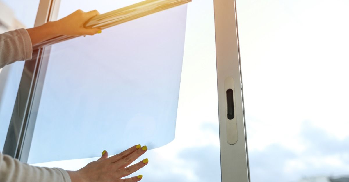 A woman holds a transparent sheet of window film to residential glass while viewing the sun shining through.