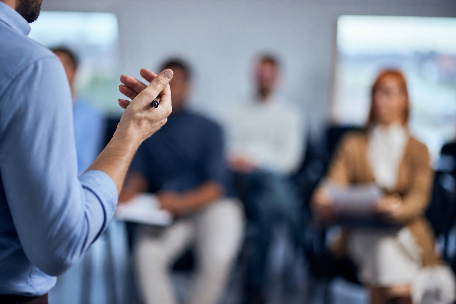 Person presenting to a seated audience in a meeting room, holding a pen in the foreground.