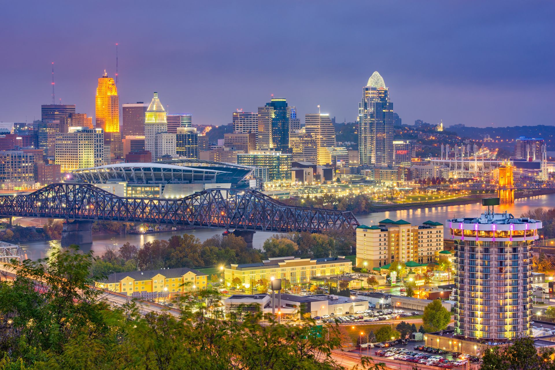 Dusk skyline of a city with lit skyscrapers, bridges, and a river in the foreground