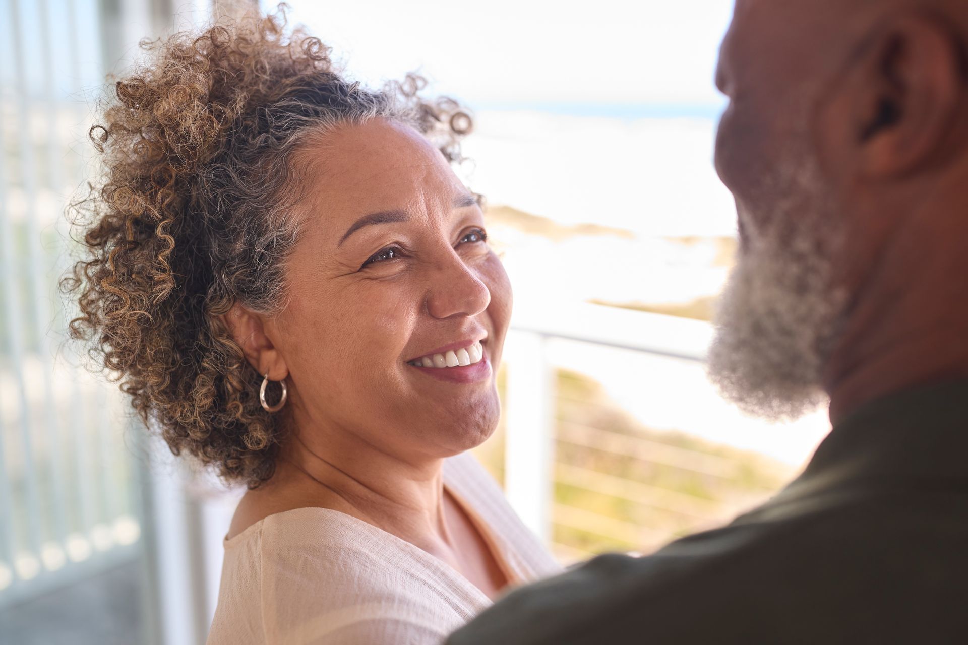 Two people smiling and talking near a bright window, with an ocean view in the background