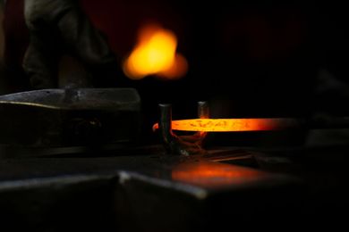 Métal orange incandescent chauffé dans une forge obscure, avec la main et les outils d'un forgeron dans l'ombre