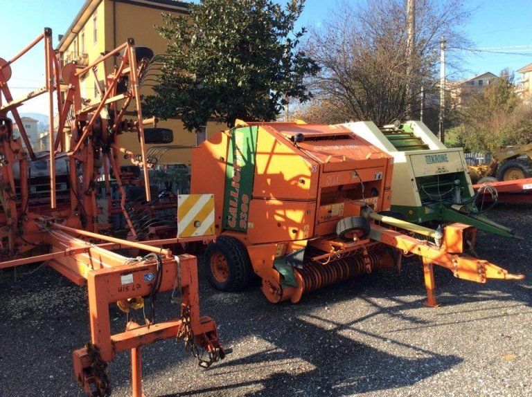 Macchinari agricoli arancioni parcheggiati all'aperto su ghiaia, con un trattore verde e altre attrezzature sullo sfondo.