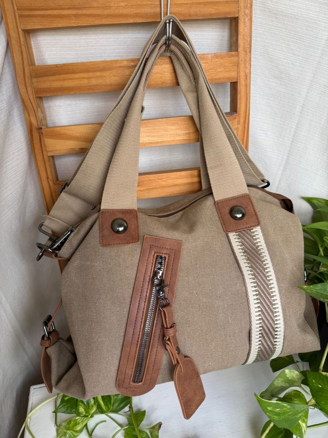 Bolso tote beige con ribete de cuero marrón y bolsillo frontal con cremallera colgado de una silla de madera.