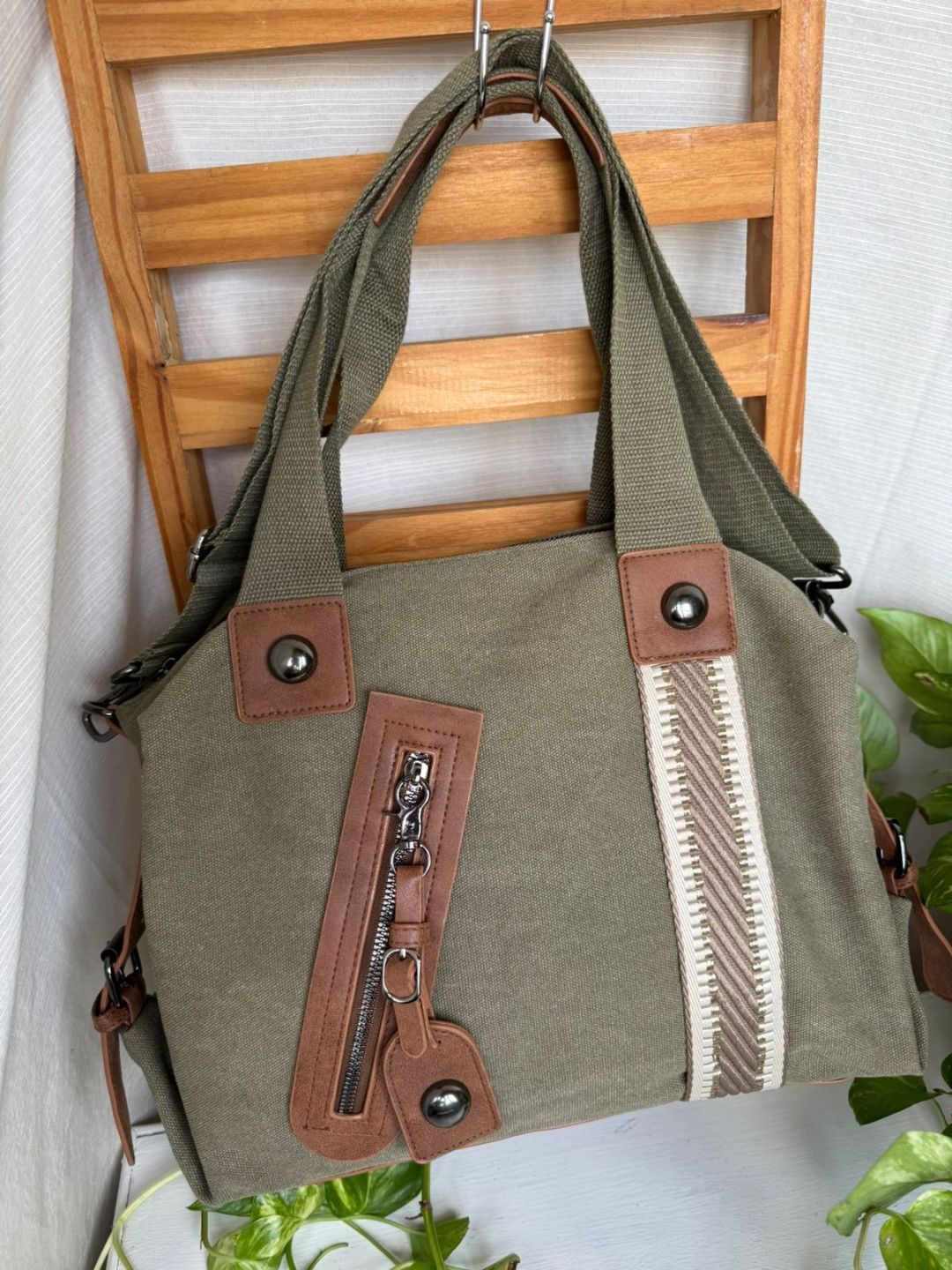 Bolso tote beige con ribete de cuero marrón y bolsillo frontal con cremallera colgado de una silla de madera.