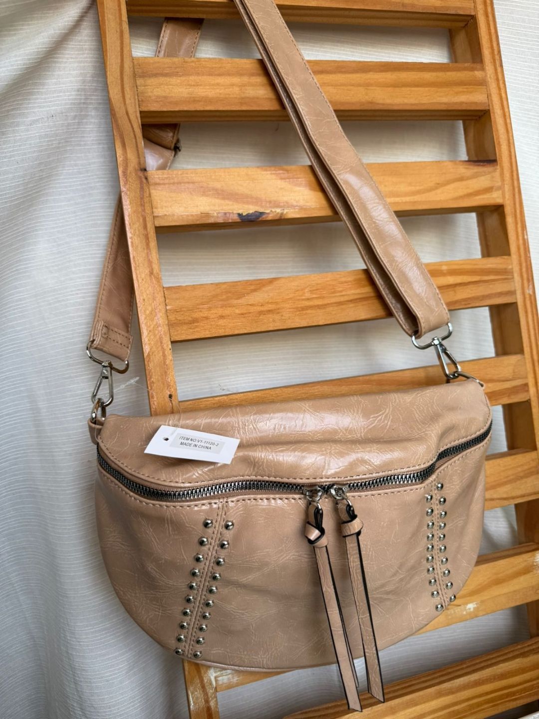 Bolso bandolera de piel color camel colgado de una escalera de madera, con tachuelas y detalle de borla.