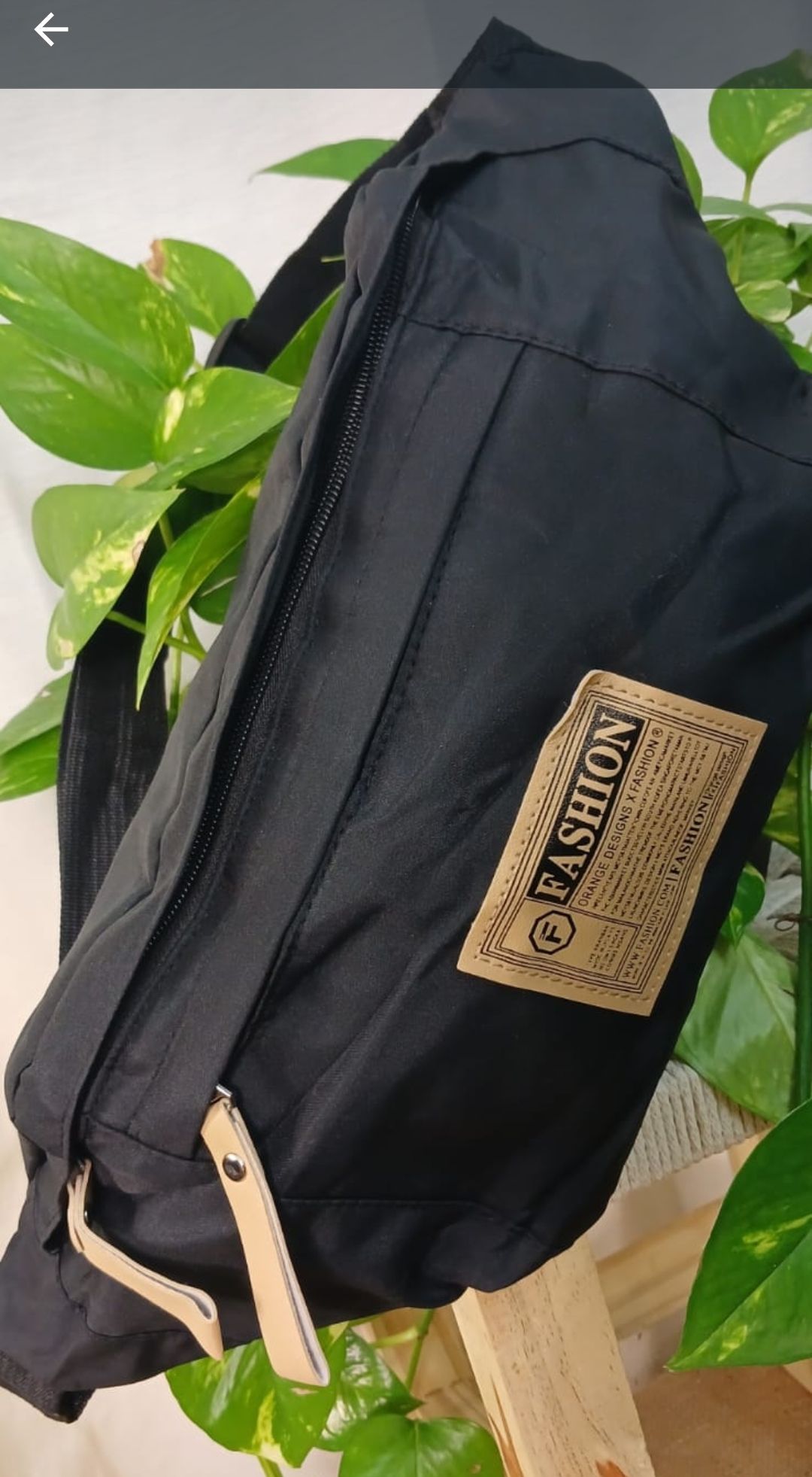 Riñonera negra con etiqueta color canela sobre un fondo blanco con estampado de plantas verdes frondosas.