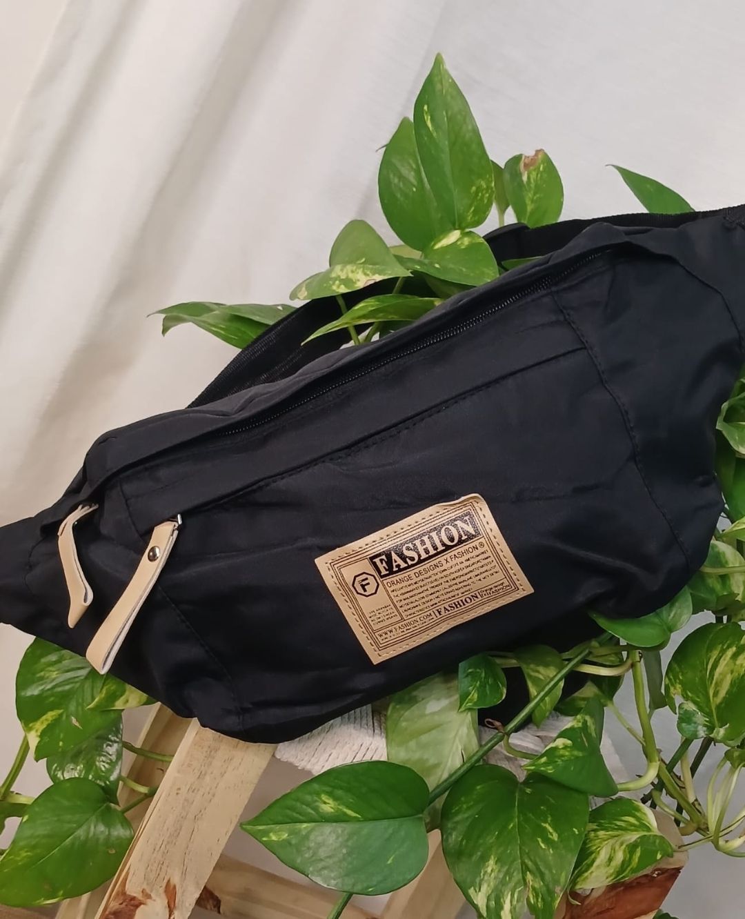 Riñonera negra con etiqueta color canela sobre un fondo blanco con estampado de plantas verdes frondosas.