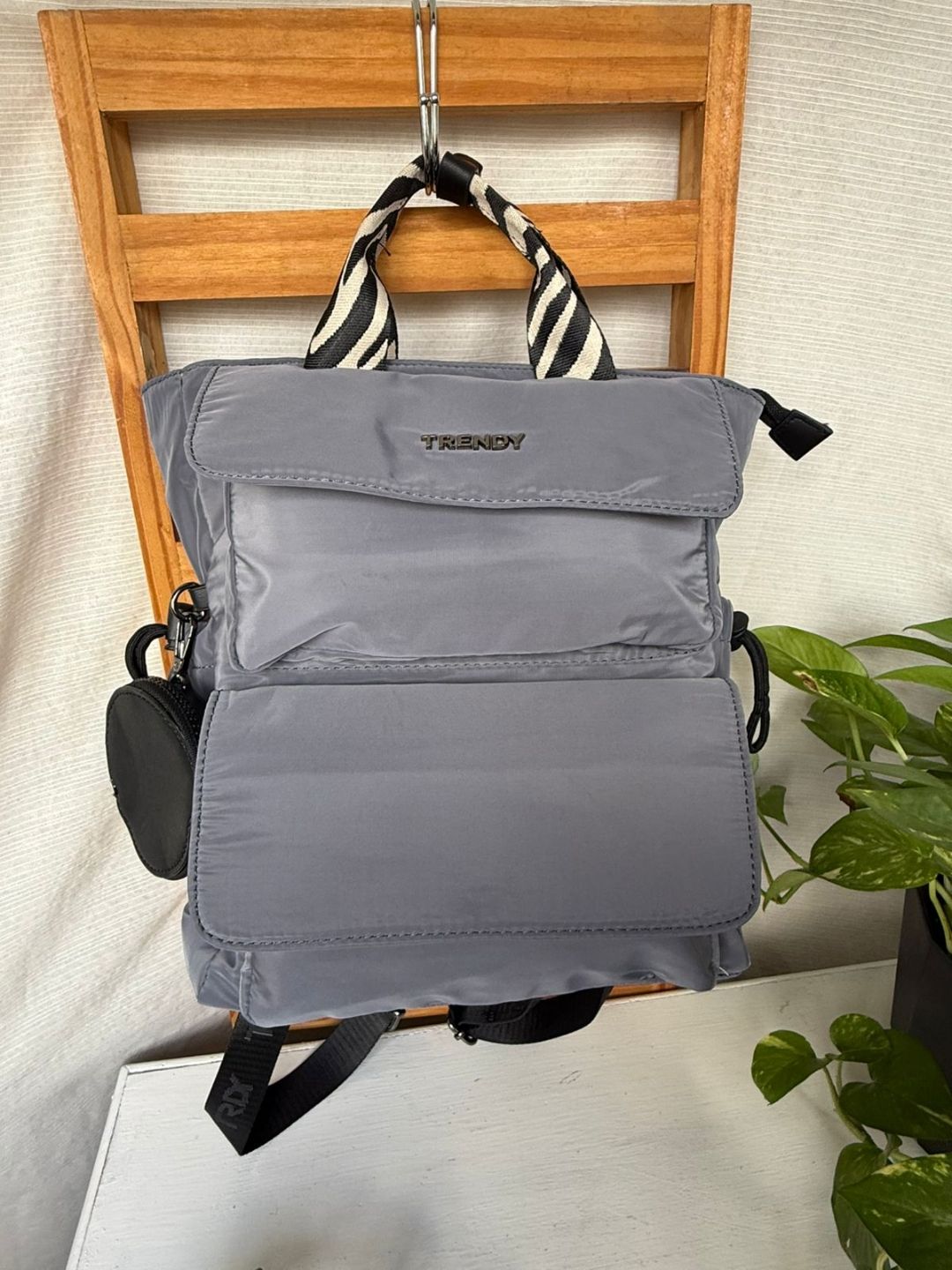 Mochila gris colgada de una silla de madera con una correa a rayas blancas y negras y un pequeño bolsillo lateral.