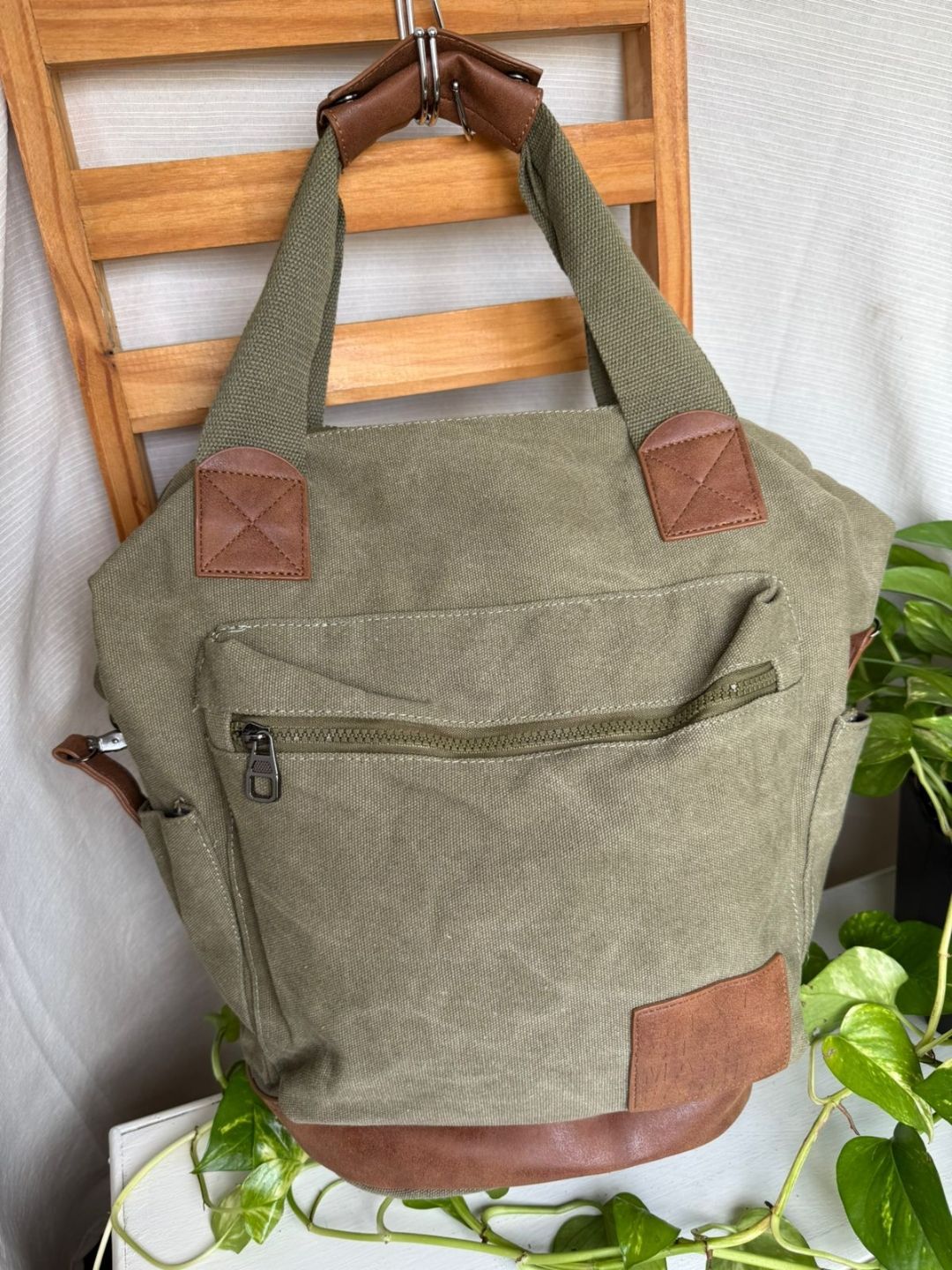 Bolso de lona verde oliva con detalles de cuero marrón colgado de un estante de madera con forma de escalera.