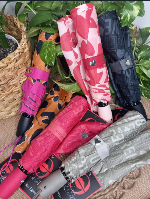 Surtido de paraguas plegados en rosa brillante, naranja, negro y gris junto a una cesta tejida y hojas verdes.